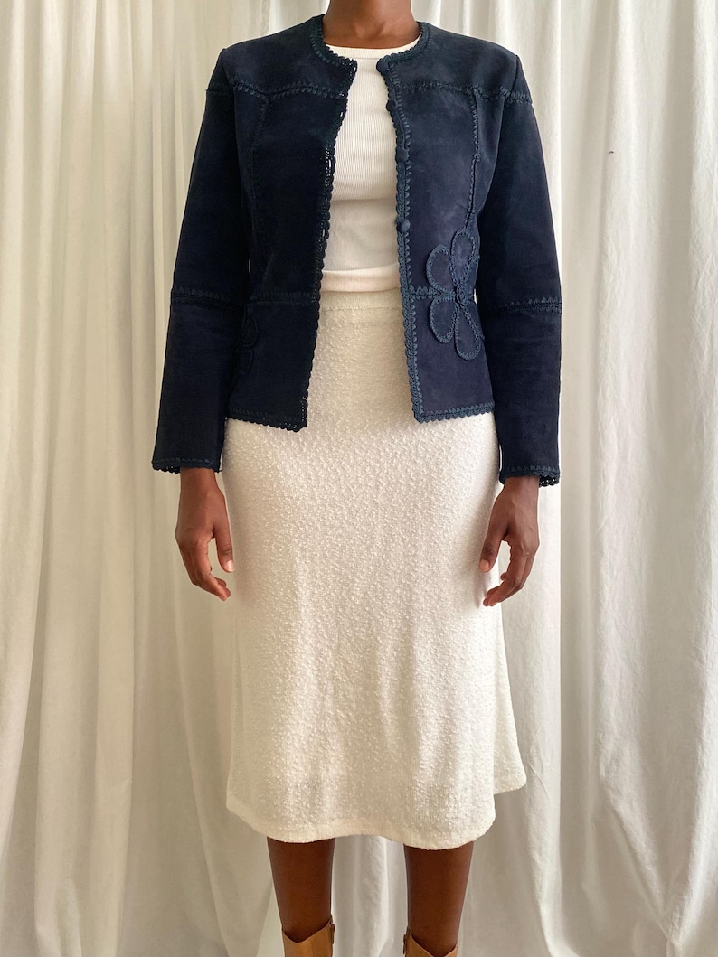 K&ouml;nnte beinhalten: Marineblaue Wildlederjacke mit floraler Applikation, kombiniert mit einem wei&szlig;en Rock. Die Jacke hat einen runden Ausschnitt und dekorative N&auml;hte. Der Rock ist ein strukturierter, knielanger Stil.