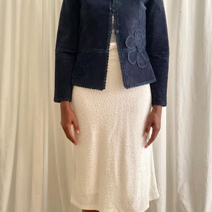 K&ouml;nnte beinhalten: Marineblaue Wildlederjacke mit floralen Applikationen. Die Jacke hat einen runden Kragen und eine Knopfleiste. Die &Auml;rmel sind lang und sie wird mit einem cremefarbenen Kleid kombiniert. Die Jacke hat einen gewellten Rand.