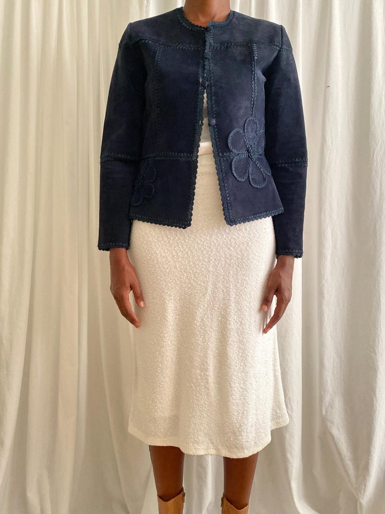 K&ouml;nnte beinhalten: Marineblaue Wildlederjacke mit floralen Applikationen. Die Jacke hat einen runden Kragen und eine Knopfleiste. Die &Auml;rmel sind lang und sie wird mit einem cremefarbenen Kleid kombiniert. Die Jacke hat einen gewellten Rand.