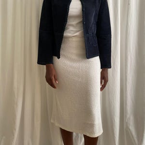 K&ouml;nnte beinhalten: Eine marineblaue Wildlederjacke mit floralem Design auf der Vorderseite, kombiniert mit einem wei&szlig;en Rippen-Top und einem wei&szlig;en strukturierten Rock. Das Outfit wird durch braune Stiefel erg&auml;nzt, wodurch ein koordiniertes Ensemble entsteht.