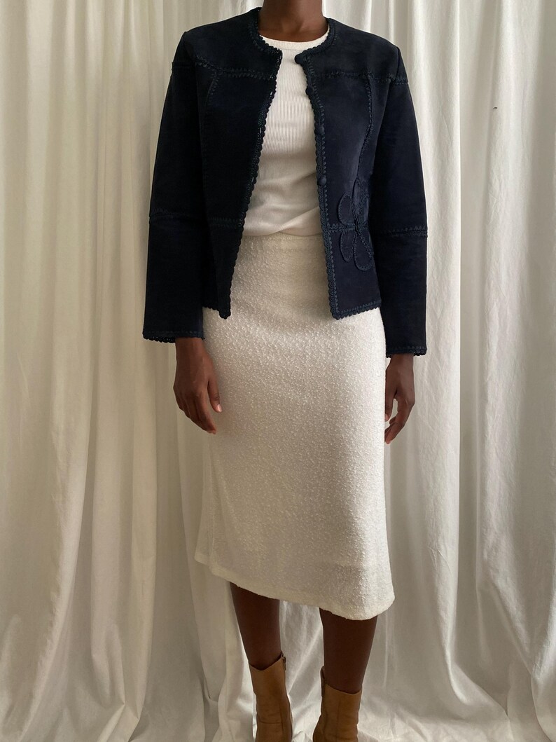 K&ouml;nnte beinhalten: Eine marineblaue Wildlederjacke mit floralem Design auf der Vorderseite, kombiniert mit einem wei&szlig;en Rippen-Top und einem wei&szlig;en strukturierten Rock. Das Outfit wird durch braune Stiefel erg&auml;nzt, wodurch ein koordiniertes Ensemble entsteht.