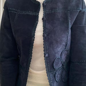 K&ouml;nnte beinhalten: Marineblaue Wildlederjacke mit floraler Applikation auf der Vorderseite. Die Jacke hat dekorative N&auml;hte entlang der N&auml;hte und um den Kragen und den Knopfverschluss. Die Jacke wird &uuml;ber einem wei&szlig;en Oberteil getragen.
