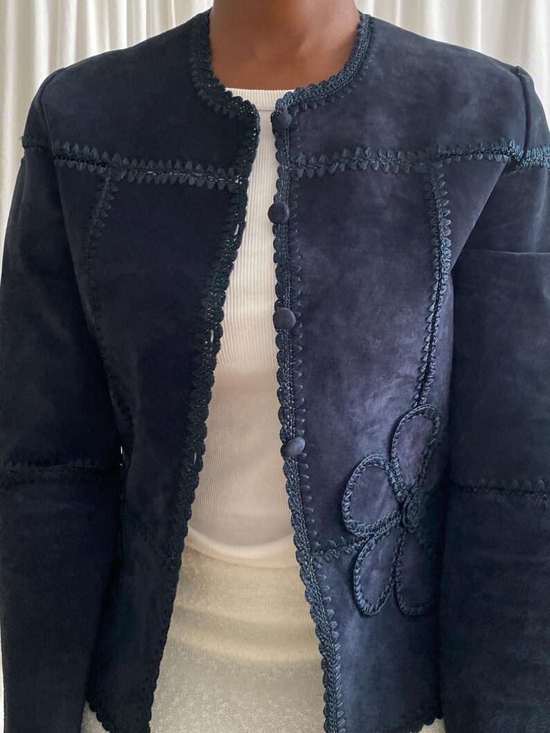 K&ouml;nnte beinhalten: Marineblaue Wildlederjacke mit floraler Applikation auf der Vorderseite. Die Jacke hat dekorative N&auml;hte entlang der N&auml;hte und um den Kragen und den Knopfverschluss. Die Jacke wird &uuml;ber einem wei&szlig;en Oberteil getragen.