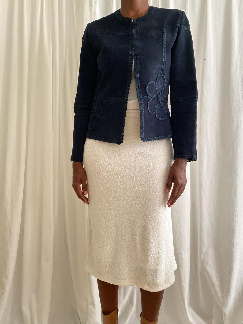 K&ouml;nnte beinhalten: Marineblaue Wildlederjacke mit floralen Applikationen und gewellten Kanten. Die Jacke hat einen runden Ausschnitt und einen Knopfverschluss. Das Model tr&auml;gt einen cremefarbenen strukturierten Rock.