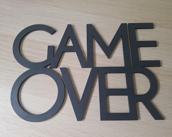 May include: A matte black wall sign featuring the words GAME OVER in a bold, sans-serif font. The letters are connected in two rows, with GAME on the top and OVER on the bottom, creating a single, flat, cutout-style decorative piece.