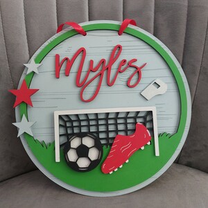 May include: A round wooden sign with a green border and a white background. The sign features a football, a football boot, a football goal, and a whistle. The name "Myles" is written in red cursive letters on the sign.