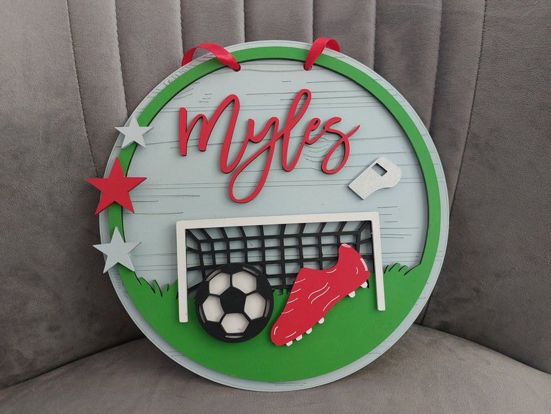 May include: A round wooden sign with a green border and a white background. The sign features a football, a football boot, a football goal, and a whistle. The name "Myles" is written in red cursive letters on the sign.
