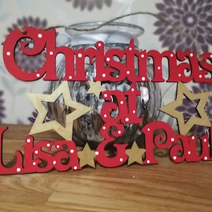 May include: Red wooden sign with white polka dots that reads "Christmas at Lisa & Paul's". The sign has gold stars and is hanging from a string.