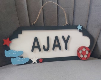 May include: A wooden sign with the name "AJAY" in black letters on a white background. The sign is decorated with blue and red stars, a red football, and two blue football boots.