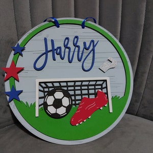May include: A round wooden sign with a green border and a white background. The sign features a football, a football boot, a whistle, and a football goal. The name "Harry" is written in blue cursive lettering on the sign.