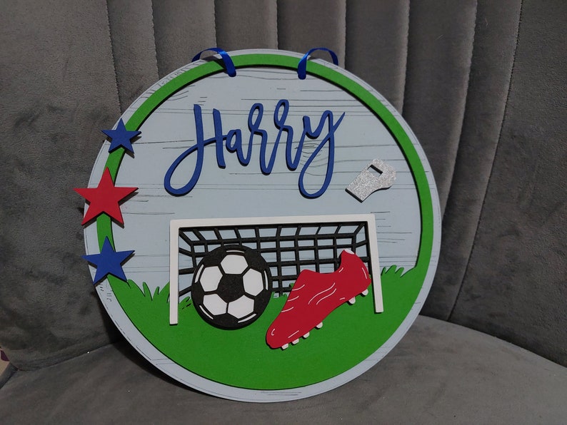May include: A round wooden sign with a green border and a white background. The sign features a football, a football boot, a whistle, and a football goal. The name "Harry" is written in blue cursive lettering on the sign.