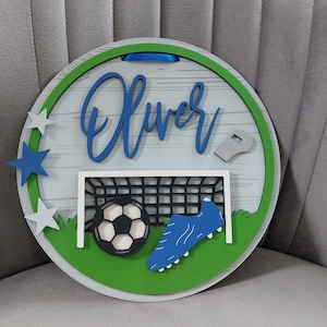 May include: A round wooden sign with a green and white border. The sign features a football, a football boot, a goal, and a whistle. The name "Oliver" is written in blue cursive lettering on the sign.