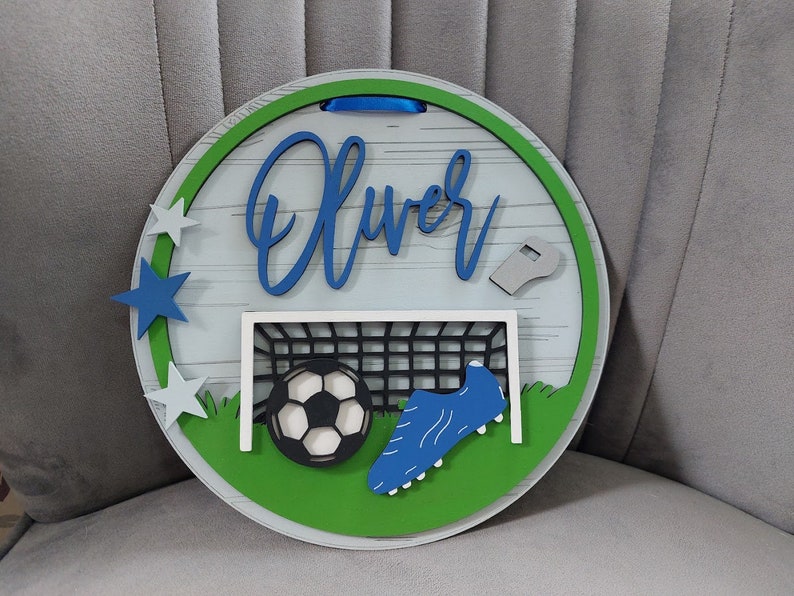 May include: A round wooden sign with a green and white border. The sign features a football, a football boot, a goal, and a whistle. The name "Oliver" is written in blue cursive lettering on the sign.