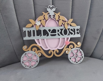 May include: A decorative wooden carriage in pink, silver, and gold, with the name "TILLY ROSE" across the center. The carriage features heart-shaped wheels and floral accents, perfect for a nursery or child's room.