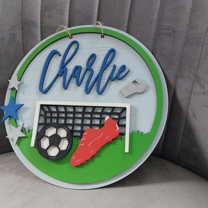 May include: A round wooden sign with a green and white football pitch design. The sign features a football, a football boot, a whistle, and a goal. The name "Charlie" is written in blue letters on the sign.