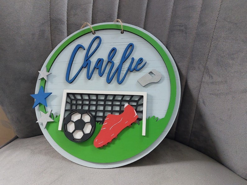 May include: A round wooden sign with a green and white football pitch design. The sign features a football, a football boot, a whistle, and a goal. The name "Charlie" is written in blue letters on the sign.