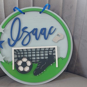 May include: A round wooden sign with a green border and a light blue background. The sign features the name "Isaac" in blue cursive lettering, a football, a football boot, a football goal, and a whistle. There are also five blue and silver stars around the border.