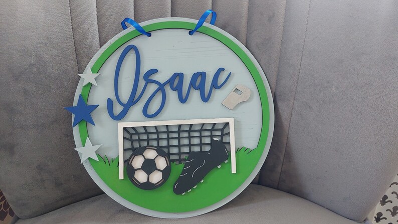 May include: A round wooden sign with a green border and a light blue background. The sign features the name "Isaac" in blue cursive lettering, a football, a football boot, a football goal, and a whistle. There are also five blue and silver stars around the border.