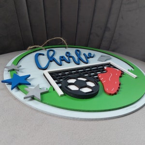 May include: A round wooden sign with a green field, a football, a goal, and a red football boot. The sign also has a blue star, two grey stars, and the name "Charlie" written in blue cursive.