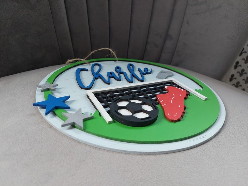 May include: A round wooden sign with a green field, a football, a goal, and a red football boot. The sign also has a blue star, two grey stars, and the name "Charlie" written in blue cursive.