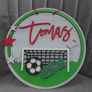 May include: A green, white and red circular wooden sign with the name "Tomas" written in red. The sign features a football, a football boot and a football goal net.