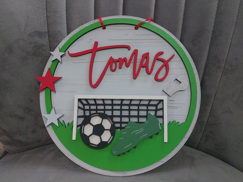 May include: A green, white and red circular wooden sign with the name "Tomas" written in red. The sign features a football, a football boot and a football goal net.