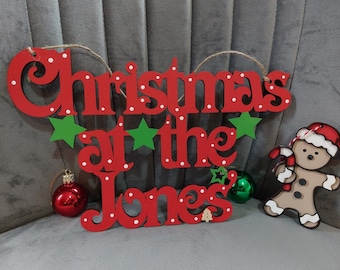 May include: A red wooden sign with white polka dots featuring the text "Christmas at the Jones" in a cursive font. The sign is decorated with green stars, a small wooden tree, and a gingerbread person ornament holding a candy cane.
