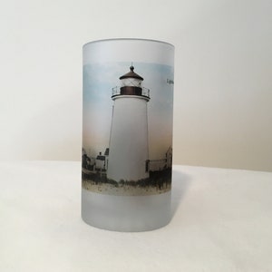 May include: A frosted glass mug with a vintage image of a lighthouse printed on it. The lighthouse is white with a brown roof and is surrounded by a sandy beach and a blue sky.