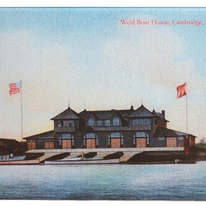 May include: A vintage postcard depicting the Weld Boat House in Cambridge, MA. The building is a large, wooden structure with a gabled roof and multiple windows. There are two flags flying in the breeze, one American and one red. The boat house is situated on a body of water with boats docked in front of it.