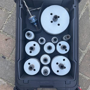May include: A black toolbox containing various hole saw blades and a drill bit. The blades are silver and circular, with different diameters. The drill bit is silver and blue. The toolbox has a clear lid and a red catch.