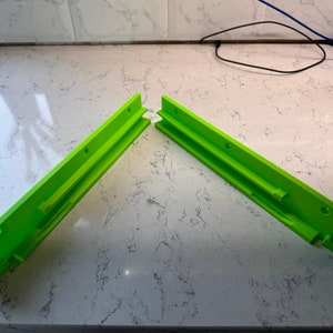 Puede incluir: Dos soportes de pared de plástico verde lima con múltiples agujeros para tornillos. Los soportes están diseñados para fijarse a una pared y tienen una ranura para una estantería u otro objeto que se pueda fijar.