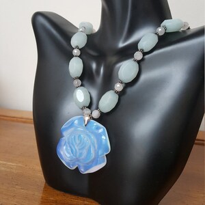 May include: A light blue iridescent glass rose pendant hangs from a necklace made of light blue and gray oval beads.