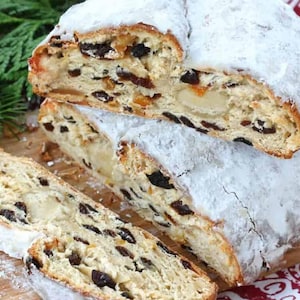 May include: A close-up of a sliced stollen, a traditional German Christmas bread, dusted with powdered sugar and filled with dried fruits and nuts. The bread is on a wooden cutting board.