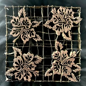 May include: A square, wooden batik stamp with a grid pattern. Four floral designs are arranged in a square, each featuring a detailed flower and leaves. The stamp is made of light brown wood with a dark brown background.