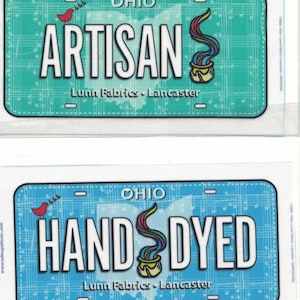 May include: Two rectangular license plate-style signs featuring a musical staff background. The top sign is teal with the text ARTISAN, and the bottom is blue with HAND DYED. Both include OHIO, a red bird, a colorful paint pot, and Lunn Fabrics Lancaster.