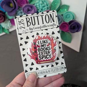 May include: A 5.7 cm button with the text "I LIKE MY BOOKS EXTRA SPICY" on a white background with a black triangle pattern. The button features a red book with flames and chili peppers. The button is by Goosefeathercrafts.