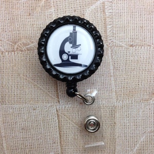 May include: Black and white microscope image on a black and white bottle cap badge reel. The badge reel has a clear plastic strap and a silver metal clip.