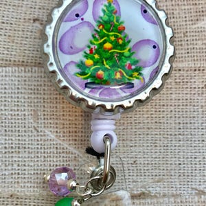 May include: A silver retractable badge holder with a purple and white Christmas tree design. The holder has a clear plastic cover and a silver chain with green and purple beads.