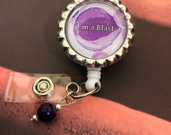 Puede incluir: Un carrete de insignia plateado con un encanto redondo morado y blanco que dice "I'm a Blast". El encanto tiene una imagen microscópica de una célula. Una cuenta morada cuelga del carrete.