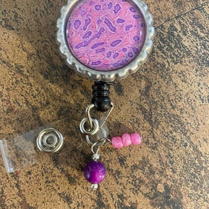 May include: A silver badge reel with a purple and pink microscope slide image on the front. The reel has a clear plastic badge holder and a purple bead with pink beads on a chain.