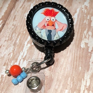 May include: A black retractable badge holder with a round, black and white button featuring a cartoon image of a red-haired character with a surprised expression. The button is attached to a clear plastic badge holder with a silver metal clip and a red and blue beaded charm.