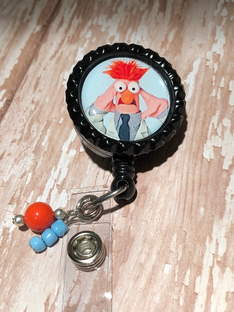 May include: A black retractable badge holder with a round, black and white button featuring a cartoon image of a red-haired character with a surprised expression. The button is attached to a clear plastic badge holder with a silver metal clip and a red and blue beaded charm.