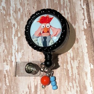 May include: A black retractable badge holder with a round, black and white button featuring a cartoon image of a red-haired character with wide eyes and a surprised expression. The button is attached to a black retractable cord and a clear plastic badge holder. The badge holder has a silver metal clip and a silver metal ring with a red bead and blue beads attached.