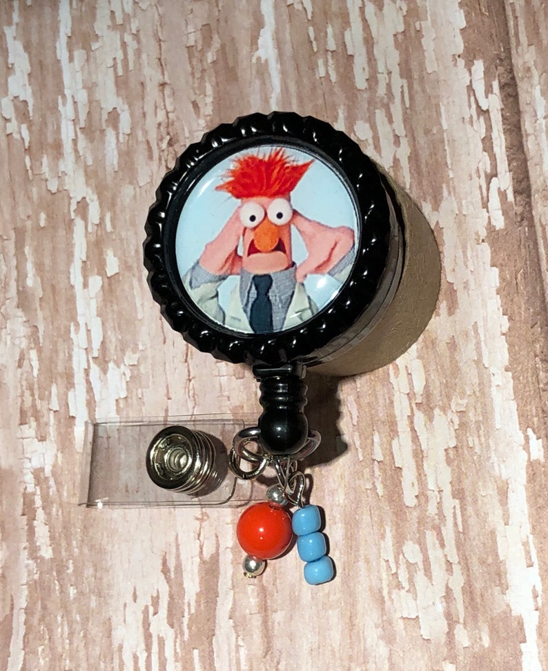 May include: A black retractable badge holder with a round, black and white button featuring a cartoon image of a red-haired character with wide eyes and a surprised expression. The button is attached to a black retractable cord and a clear plastic badge holder. The badge holder has a silver metal clip and a silver metal ring with a red bead and blue beads attached.