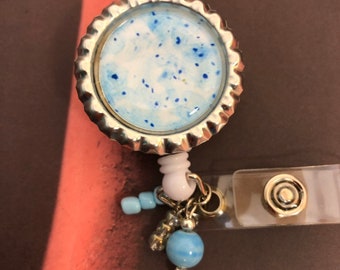 Puede incluir: Un portagafete retráctil plateado con un círculo estampado en azul y blanco en el centro. El portagafete tiene una correa de plástico transparente y un clip plateado. El portagafete está decorado con cuentas azules y plateadas.