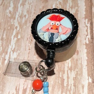 May include: A black and silver retractable badge holder with a round, black button featuring a photo of a red-haired Muppet character with a surprised expression. The button has a white background and the character is wearing a white lab coat and a blue tie. The badge holder has a clear plastic strap and a silver bead with a red bead and blue beads attached.