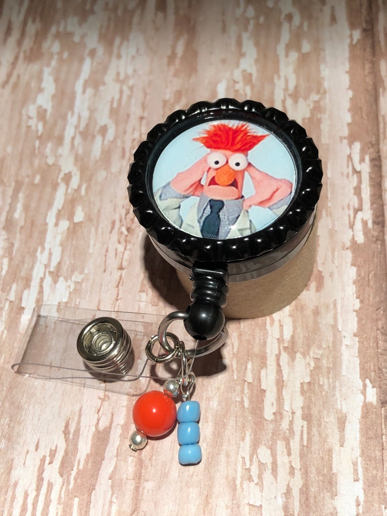 May include: A black and silver retractable badge holder with a round, black button featuring a photo of a red-haired Muppet character with a surprised expression. The button has a white background and the character is wearing a white lab coat and a blue tie. The badge holder has a clear plastic strap and a silver bead with a red bead and blue beads attached.