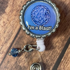 May include: A round, silver-colored badge reel with a blue and white graphic that says "I'm a Blast!" The badge reel has a clear plastic strap and a blue bead charm.