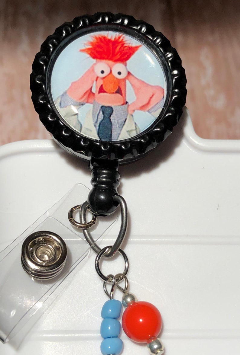May include: A black retractable badge holder with a round, black cap featuring a photo of a red-haired Muppet character with a surprised expression. The badge holder has a clear plastic clip and a silver chain with blue and red beads.