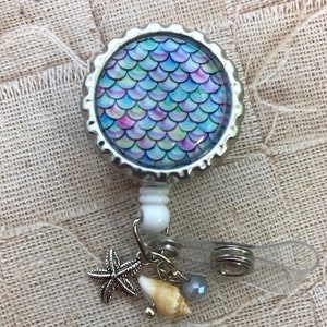 May include: A silver retractable badge holder with a mermaid scale design. The holder has a clear plastic strap and a charm with a seashell, a starfish, and a bead.